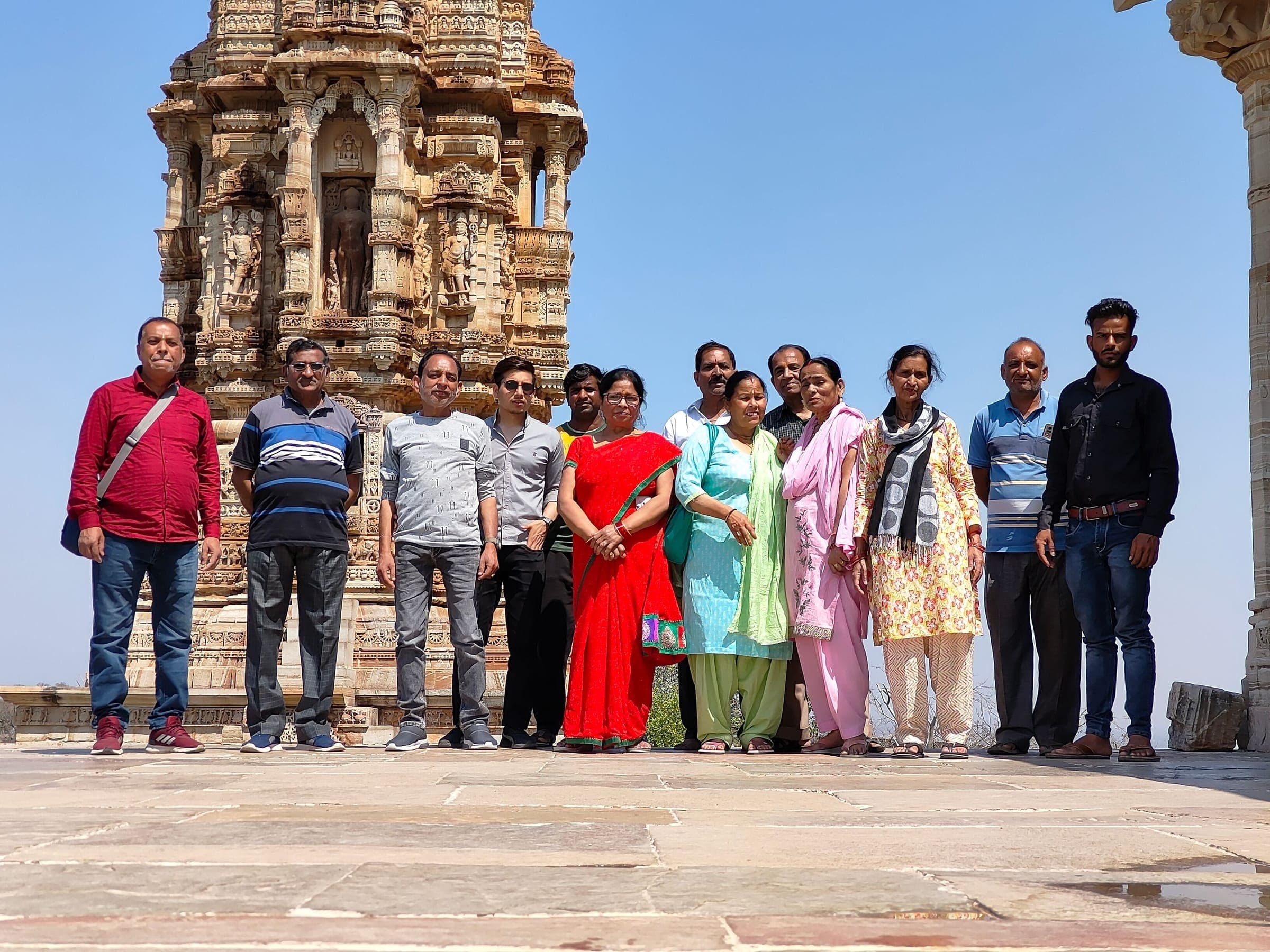 Travellers lined up on a stone terrace in front of the sculpted base of the Kirti Stambh