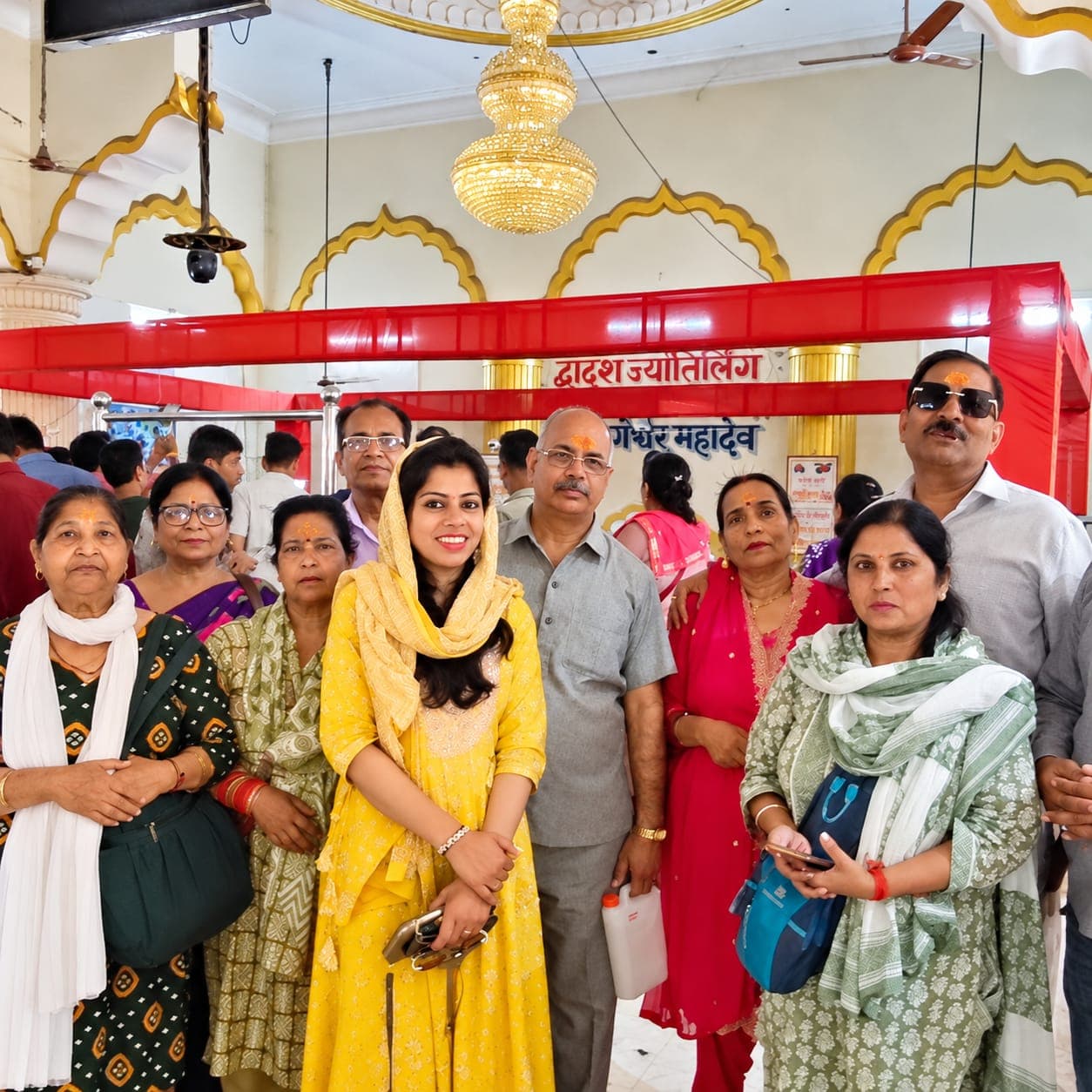 Travellers with tilak on their foreheads gathered inside a Jyotirlinga temple hall beneath a chandelier